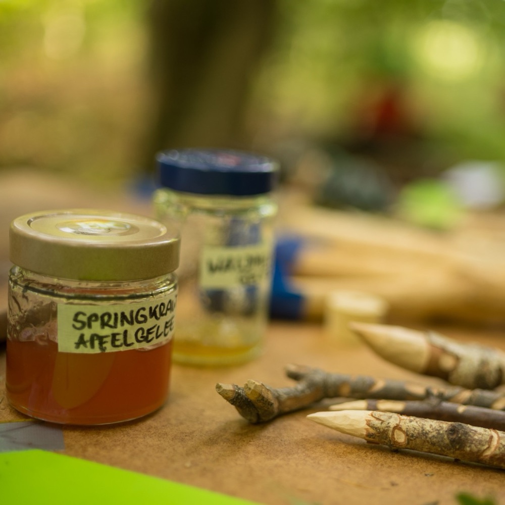 Ein Glas Springkraut-Apfelgelee neben geschnitzten Stöcken auf einem Tisch im Wald